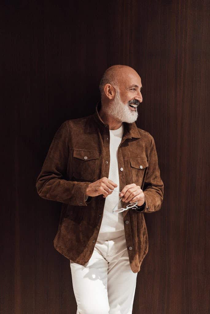 Cheerful elderly ethnic male with gray beard in fashionable outfit smiling and looking away while standing against wooden wall
